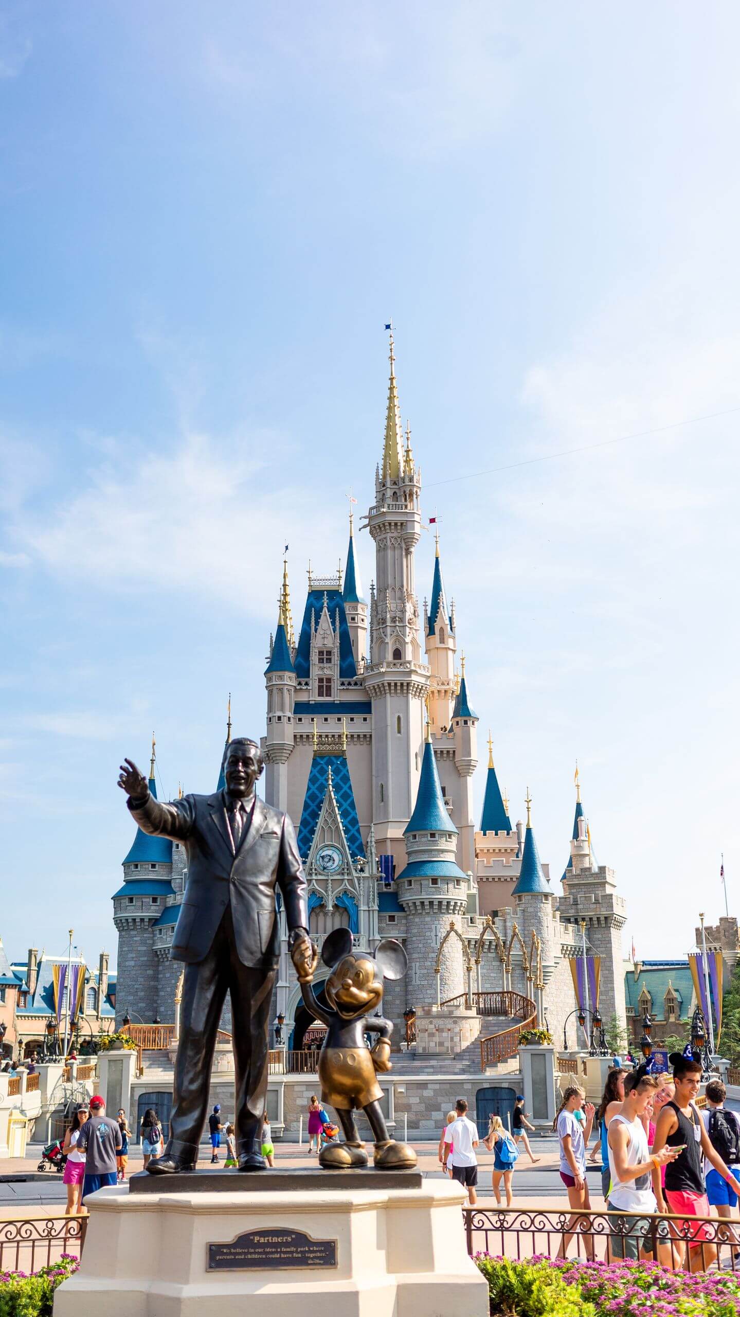 A sunny day at a famous theme park with a castle and bronze statues surrounded by tourists.